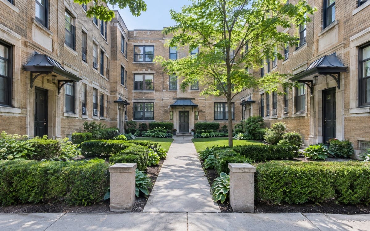A vintage courtyard apartment building, trimmed hedges lining the center walk to the front door.