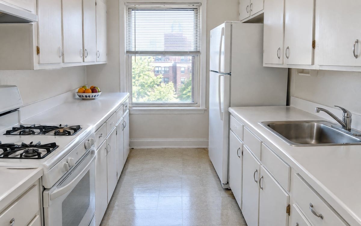 A small galley kitchen that an apartment cleaning service keeps up, white counters and a stainless sink beside a bowl of fruit.