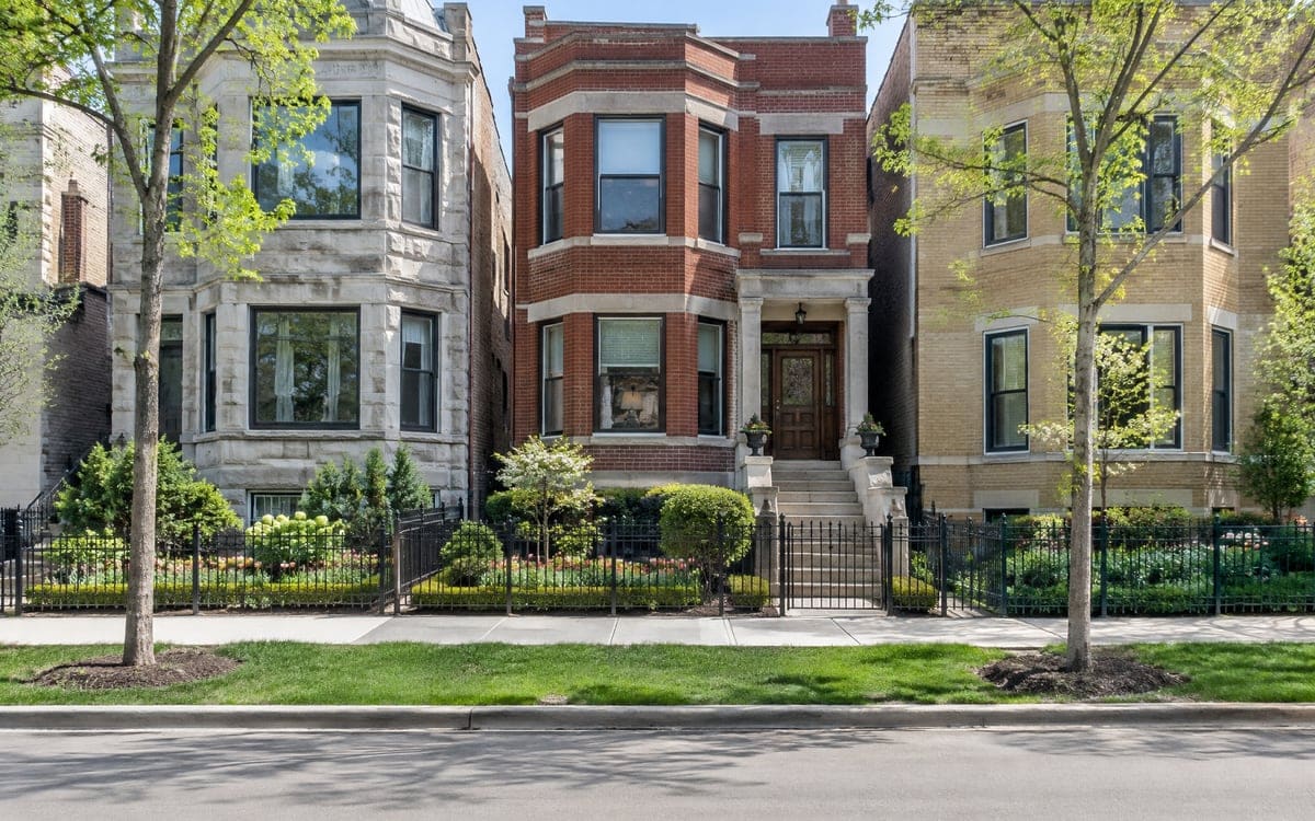 A greystone, a red brick two-flat and a yellow-brick house behind iron fences on a tree-lined block.