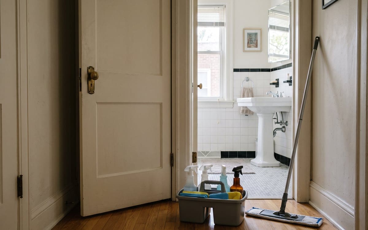 A house cleaning caddy and a flat mop wait at a bathroom doorway, a vintage pedestal sink and tile inside.
