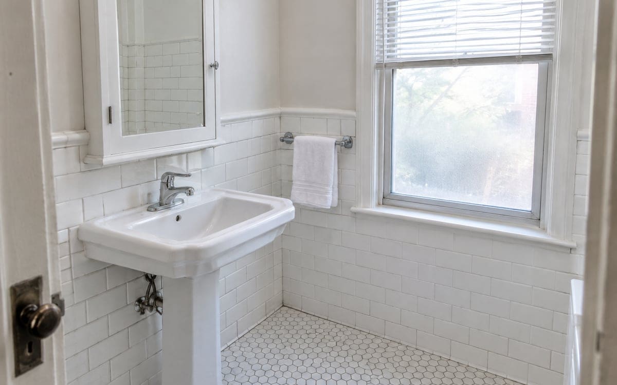 A vintage bathroom after a maid service cleaning, with a pedestal sink, white subway tile and a white hex-tile floor.